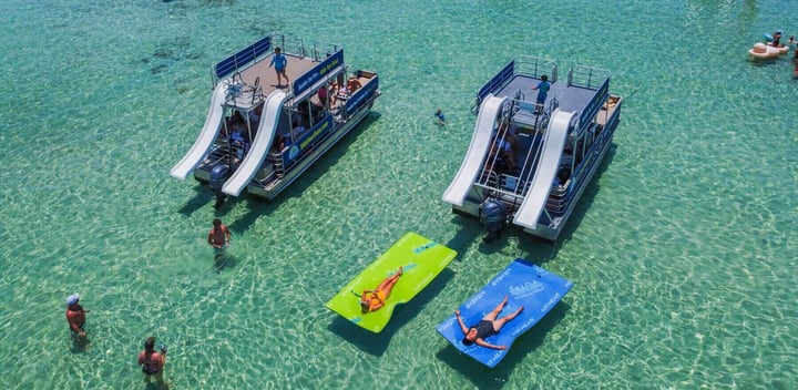 two double-decker boats anchored at Crab Island in Destin Florida next to two ladies relaxing on floating lily pad mats at Crab Island