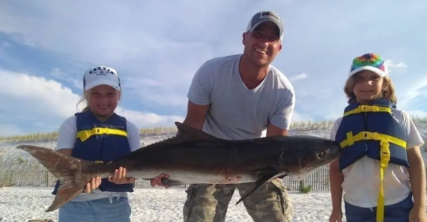 man holding a shark next to two girls on the beach